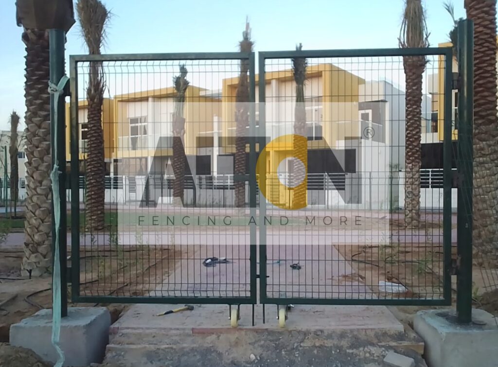 Construction site with large, grey concrete blocks placed at regular intervals, supporting a tall white metal fence. Vegetation is visible above and through the fence, with a street lamp in the background under clear skies.