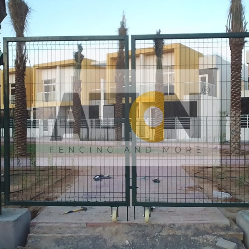 Construction site with large, grey concrete blocks placed at regular intervals, supporting a tall white metal fence. Vegetation is visible above and through the fence, with a street lamp in the background under clear skies.