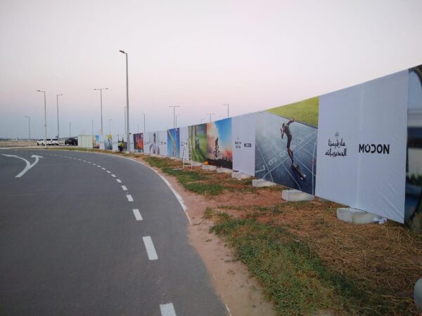 Long road with outdoor advertising fencing displaying various banners, including Modon branding, installed by Alton Fence along a curved road under clear evening skies