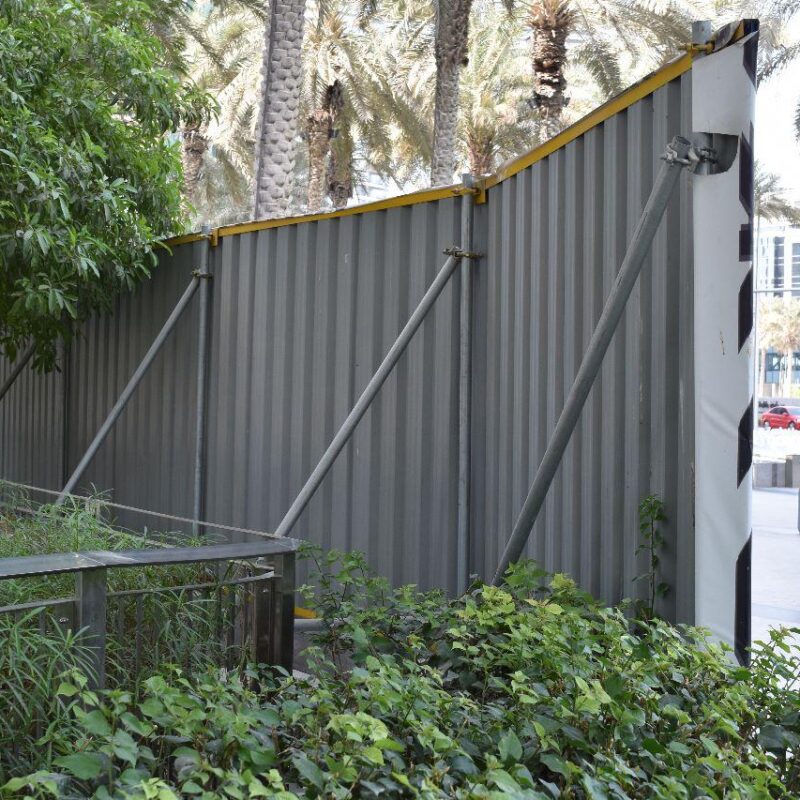 A large metal structure, possibly a fence or barrier, bent and deformed, leaning towards a walkway lined with green shrubbery and tall palm trees in the background