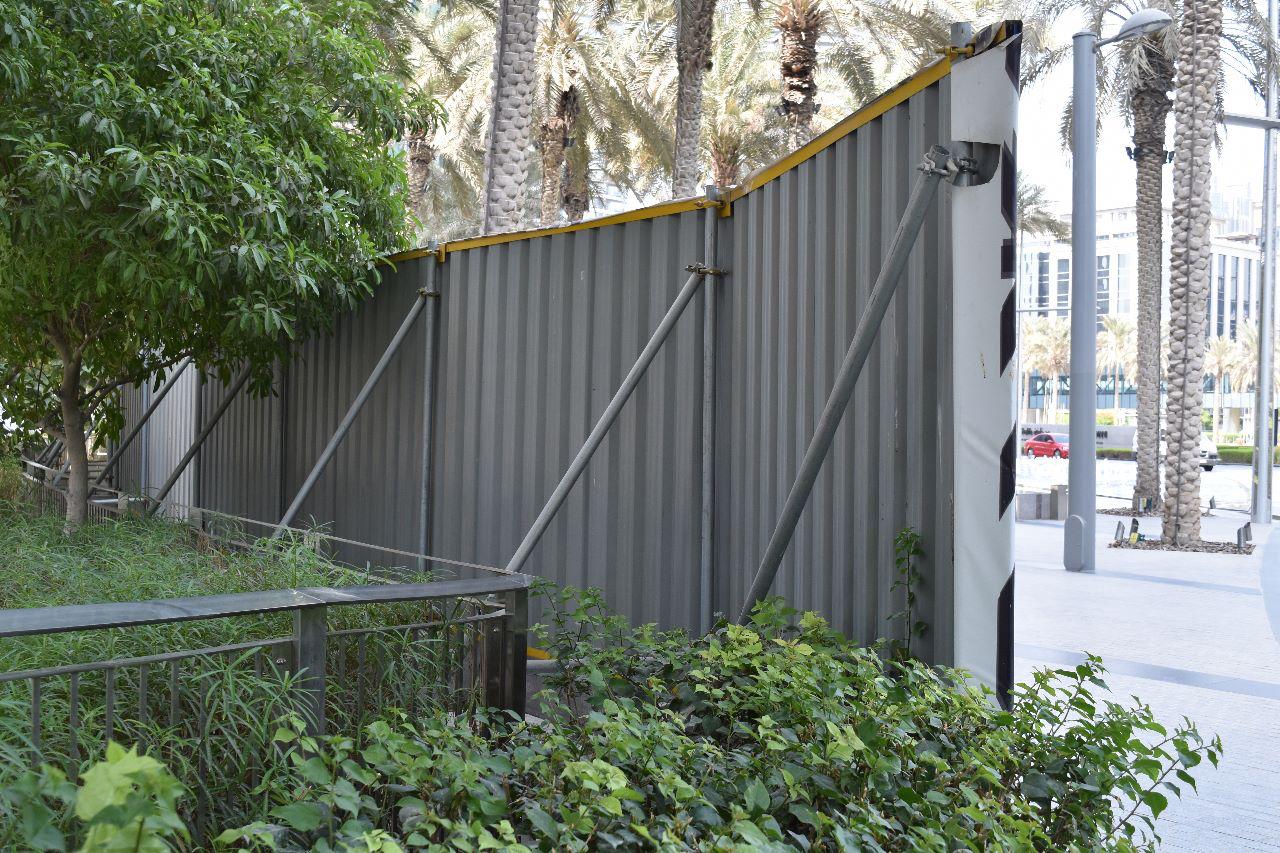 A large metal structure, possibly a fence or barrier, bent and deformed, leaning towards a walkway lined with green shrubbery and tall palm trees in the background