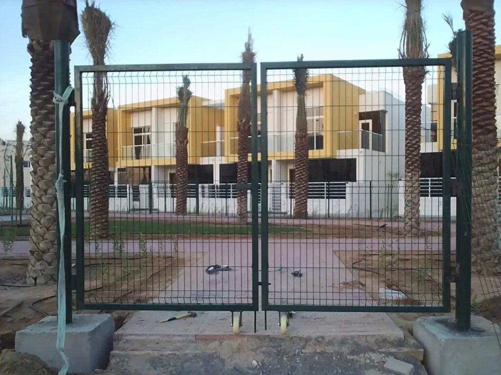 Construction site with large, grey concrete blocks placed at regular intervals, supporting a tall white metal fence. Vegetation is visible above and through the fence, with a street lamp in the background under clear skies.