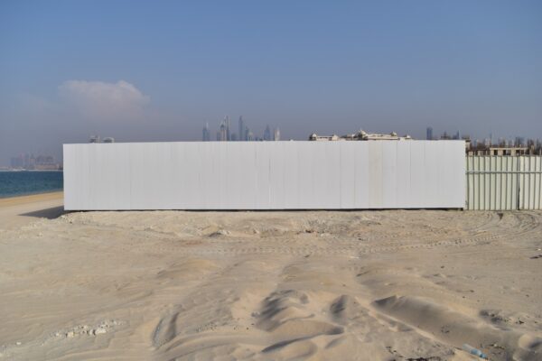 A large white barrier or wall on a sandy beach with a city skyline and tall buildings in the background under a partly cloudy sky