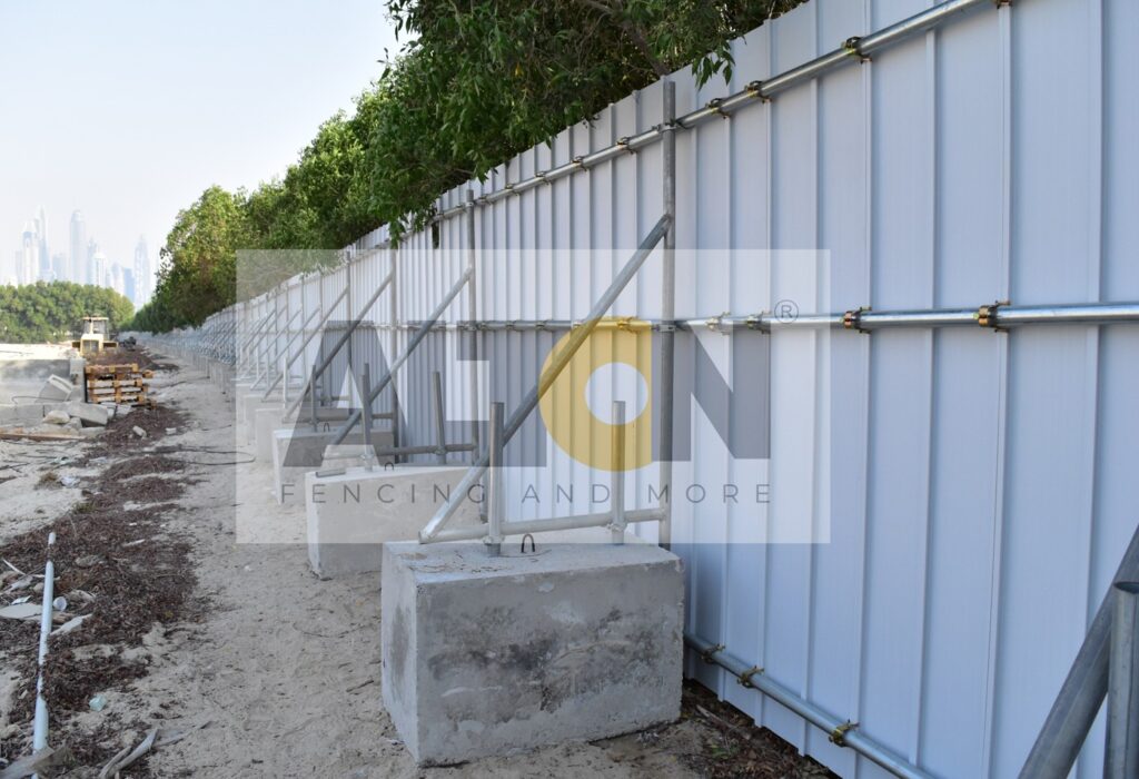 Construction site with large concrete blocks supporting metal fencing panels, with ‘BIGLAND & MORE’ watermark text across the center. Trees in the background and construction materials in the foreground.