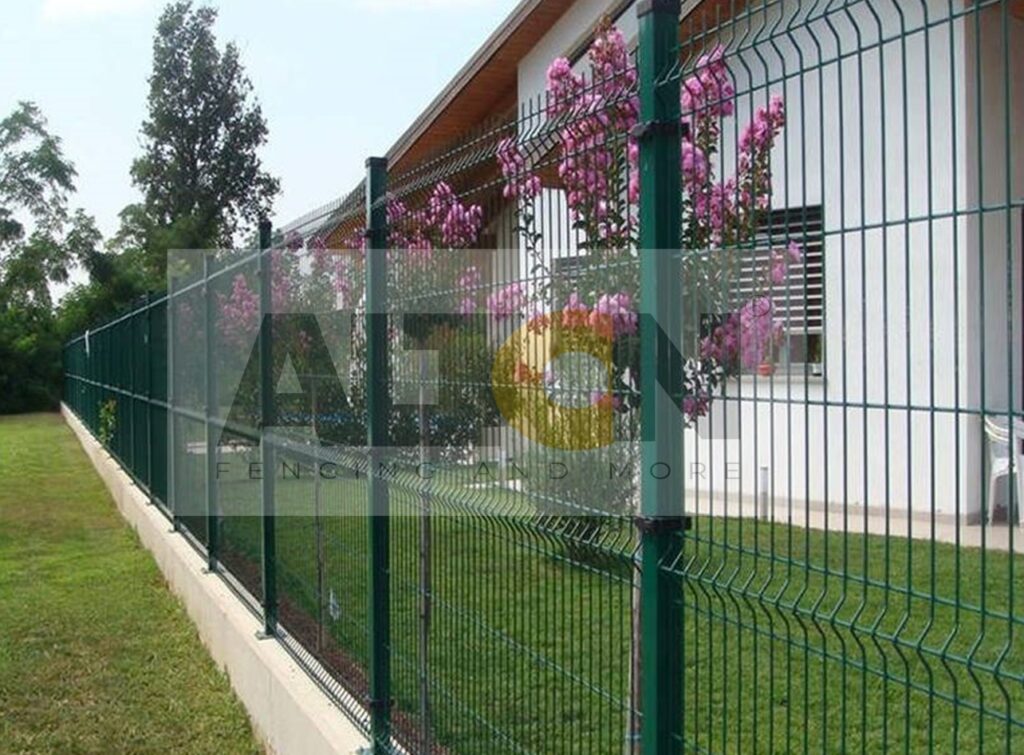 Green fence at a construction site with scattered construction materials and a white building in the background. A paved sidewalk and an individual walking away are visible on the right side under a clear, sunny sky.