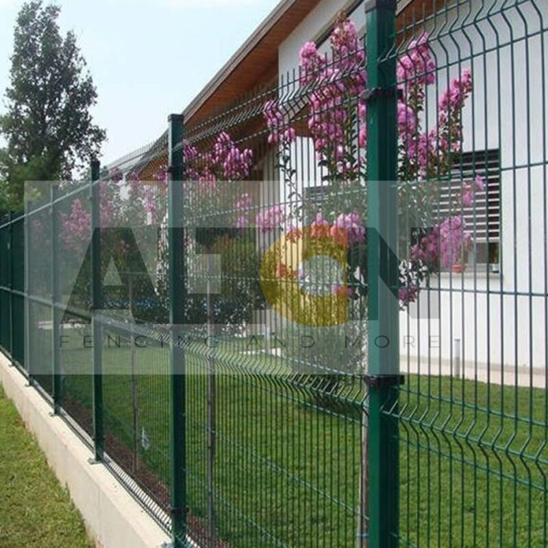 Green fence at a construction site with scattered construction materials and a white building in the background. A paved sidewalk and an individual walking away are visible on the right side under a clear, sunny sky.