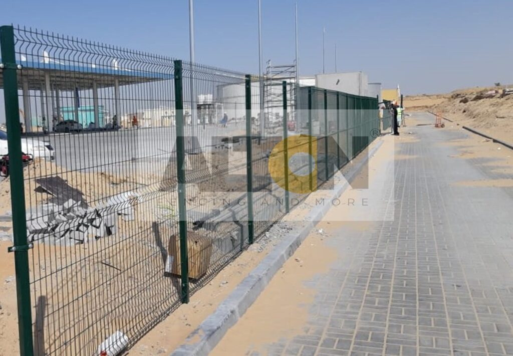 Green fence at a construction site with scattered construction materials and a white building in the background. A paved sidewalk and an individual walking away are visible on the right side under a clear, sunny sky.