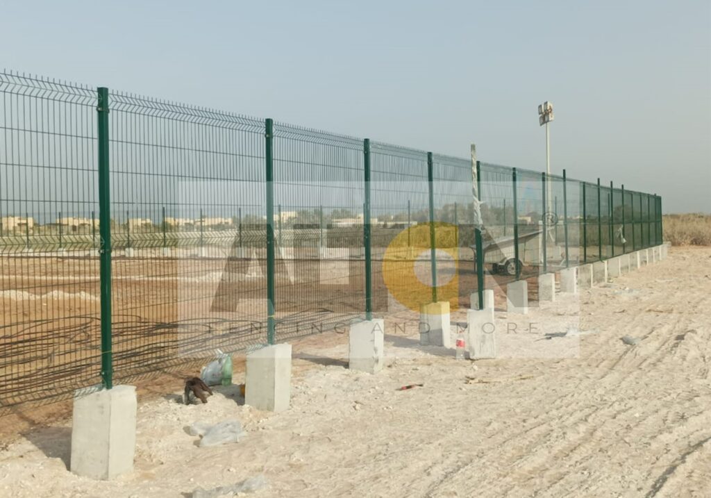 Long metal fence with green mesh running along its length, installed on a barren landscape with sparse vegetation in the background. Construction or excavation equipment is visible behind the fence, suggesting an area under development or maintenance