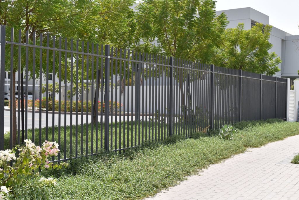 A metal fence with vertical bars situated alongside a paved walkway, with green bushes and trees behind it. The setting appears to be in an urban area with buildings in the background.