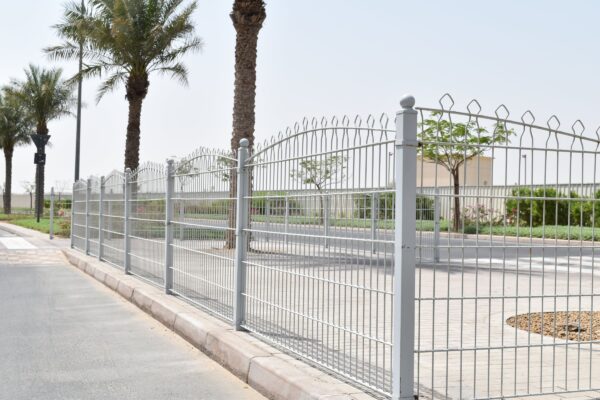 A metal fence with pointed tops along a sidewalk, with palm trees behind it and a clear sky above. The fence is likely for security and boundary definition around the property it encloses