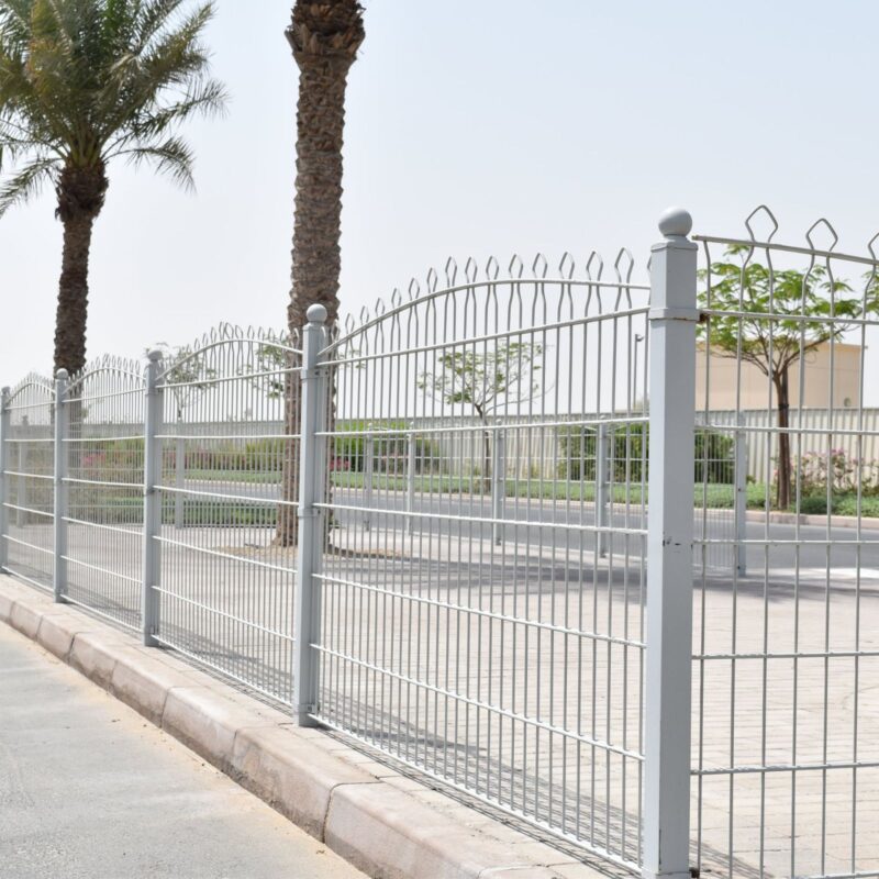 A metal fence with pointed tops along a sidewalk, with palm trees behind it and a clear sky above. The fence is likely for security and boundary definition around the property it encloses