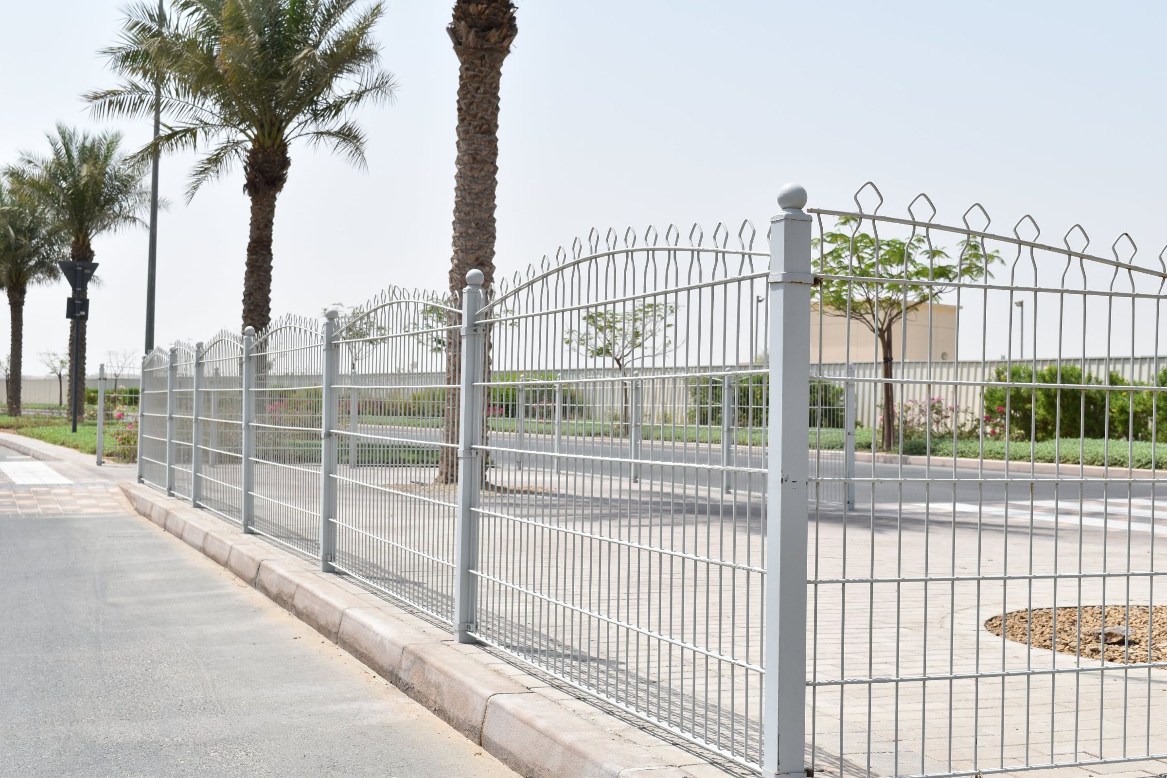 A metal fence with pointed tops along a sidewalk, with palm trees behind it and a clear sky above. The fence is likely for security and boundary definition around the property it encloses