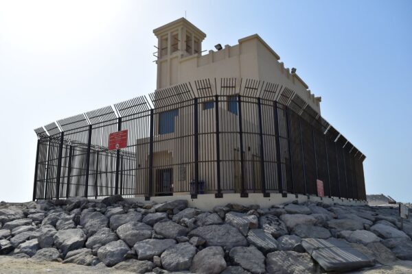 A modern building with a curved facade featuring vertical metal bars and a flat roof topped with a small tower structure, surrounded by large rocks and a clear blue sky in the background.