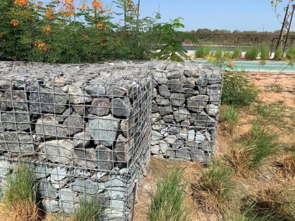 Two large gabion baskets filled with rocks and stones, with a cast aluminium fence installed on top. The fence has vertical bars and horizontal supports, situated in a well-maintained garden area with green plants.