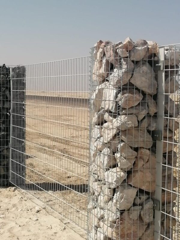 A cast aluminium fence with vertical bars and horizontal supports, installed on sandy terrain with clear skies in the background. Adjacent to the fence is a gabion wall filled with irregularly shaped stones.