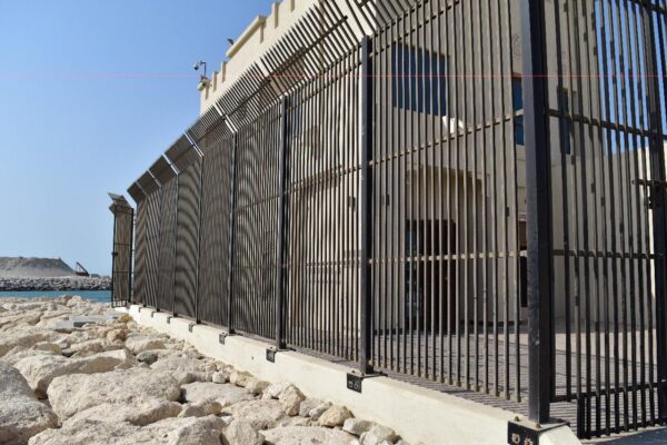 A black metal fence with vertical bars and decorative elements at the top, running parallel to a rocky shoreline with a calm sea and clear sky in the background.