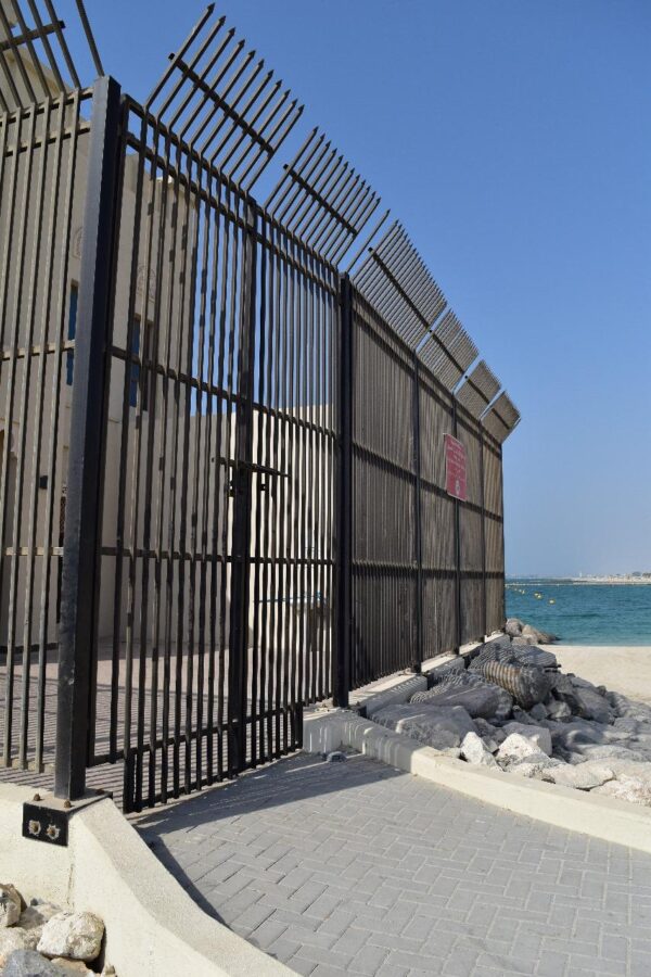 A tall, vertical metal fence with horizontal slats overlooking a coastal area, with a paved area and natural rocks in the foreground under a clear blue sky.